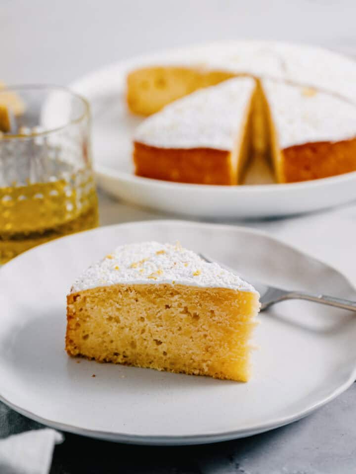 A slice of lemon ricotta cake made with olive oil on a white plate, with the whole cake and a glass of olive oil in the background.