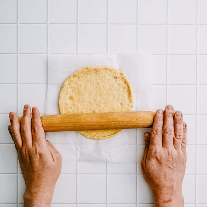 Hands rolling out chickpea flatbread dough with a rolling pin.