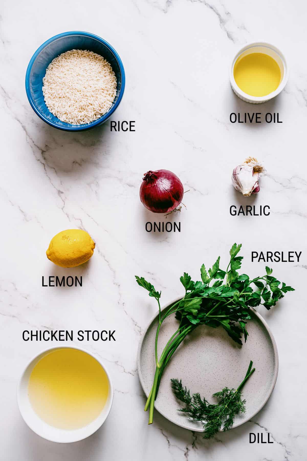 A marble worktop with bowls and plates holding labelled ingredients: rice, olive oil, chicken stock, a red onion, garlic cloves, a lemon, parsley, and dill.