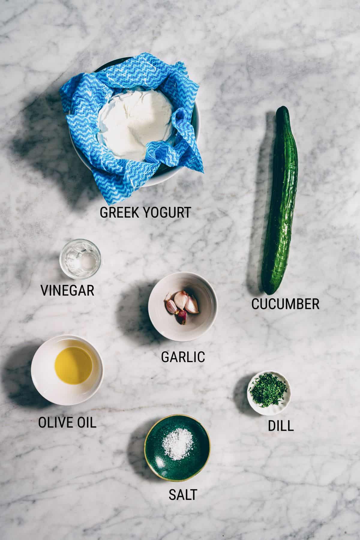 Overhead view of ingredients labelled for tzatziki: a bowl of Greek yoghurt, a whole cucumber, small bowls of vinegar, olive oil, salt, chopped dill, and two garlic cloves on a marble surface.