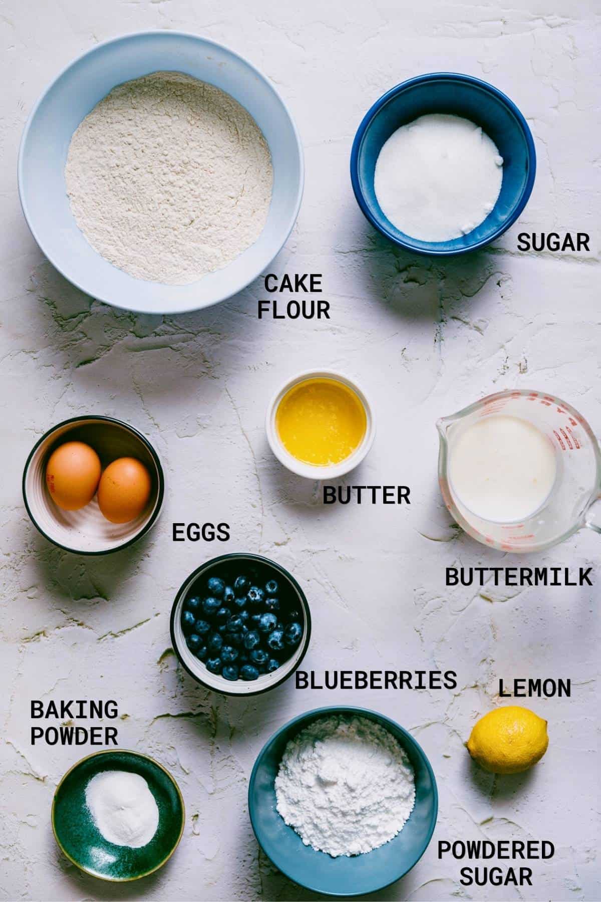 A flat lay of baking ingredients in bowls labelled as cake flour, sugar, butter, buttermilk, eggs, blueberries, baking powder, lemon, and icing sugar-everything needed to make delicious baked blueberry cake doughnuts-arranged on a white surface.