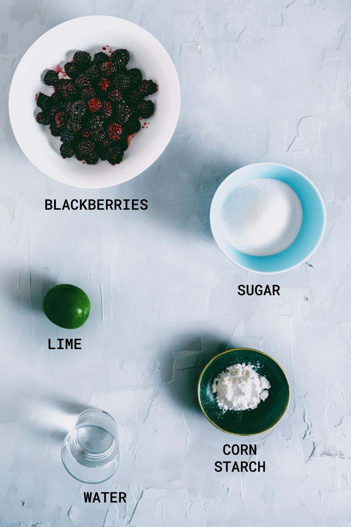 A flat lay of five ingredients for blackberry jam on a light surface: a bowl of blackberries, sugar, a whole lime, cornflour, and water. Each is labelled with text.