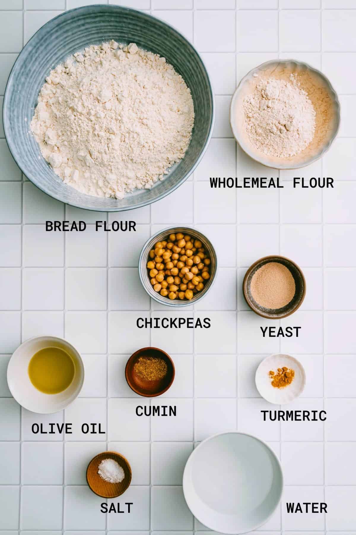 A top-down view of labelled baking ingredients on a white tiled surface: bread flour, wholemeal flour, chickpeas, yeast, olive oil, cumin, turmeric, salt, and water arranged in bowls and cups.
