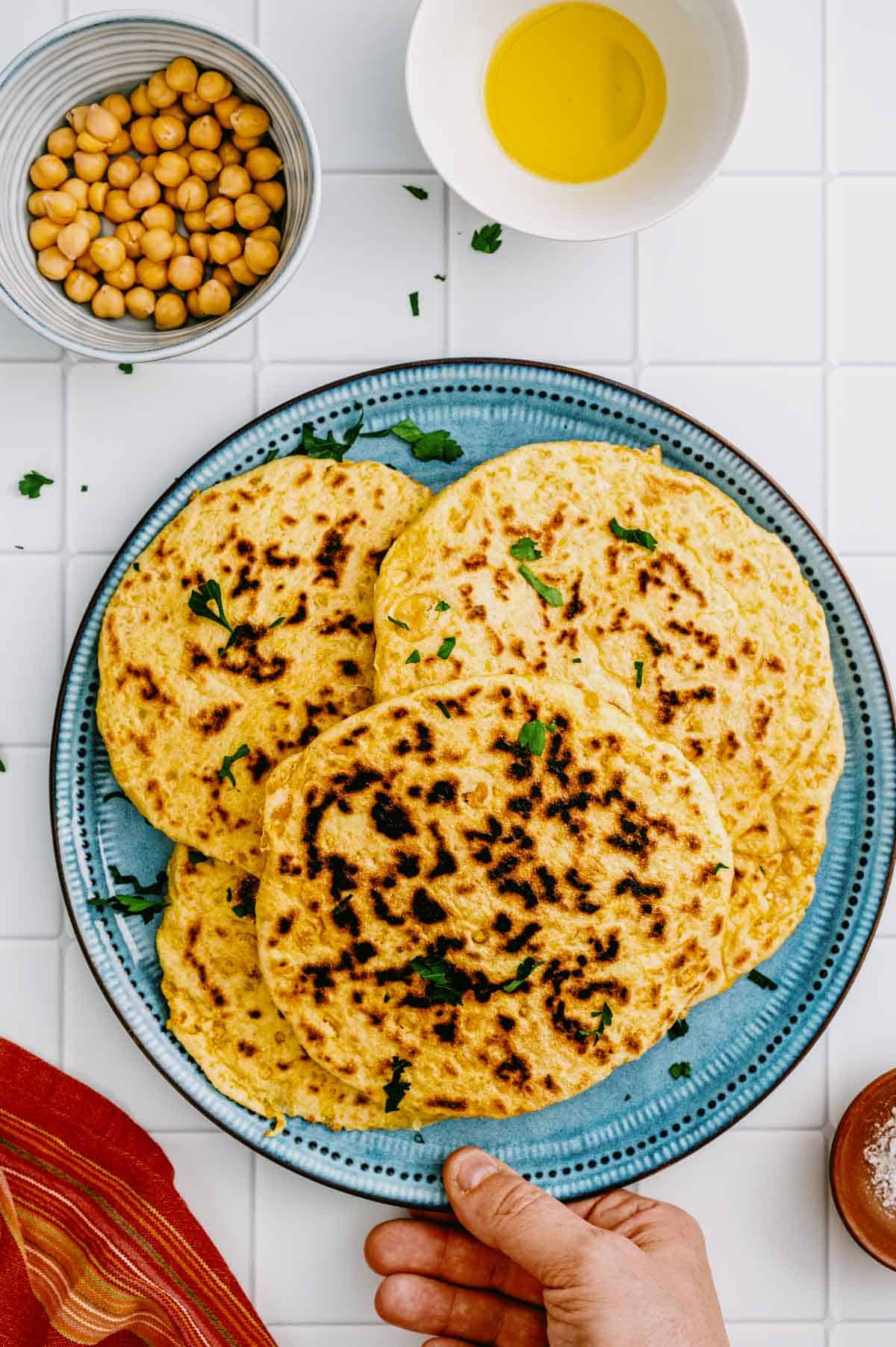 A hand holding a blue plate with several round, golden flatbreads garnished with chopped herbs, surrounded by a bowl of chickpeas, a bowl of oil, and a red-striped serviette on a white tiled surface.