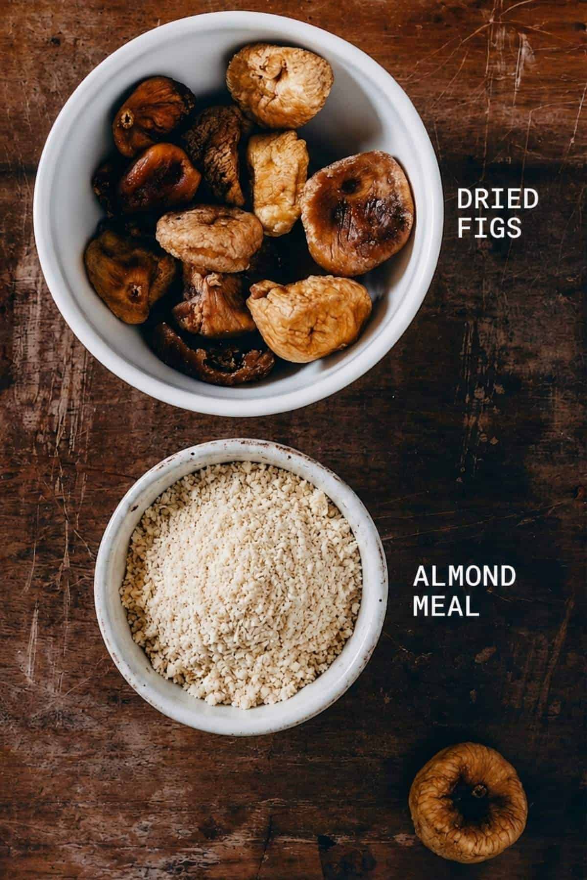 Two bowls on a rustic wooden surface: the top bowl contains dried figs, labelled DRIED FIGS, and the bottom bowl holds ground almonds, labelled GROUND ALMONDS. A single dried fig sits beside the bowls.