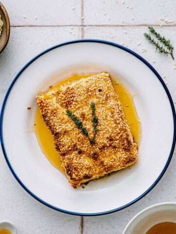 A rectangular piece of sesame-crusted feta drizzled with honey, garnished with two sprigs of herbs, served on a white plate with a blue rim. Bowls of sesame seeds and honey sit nearby on a white tiled surface.