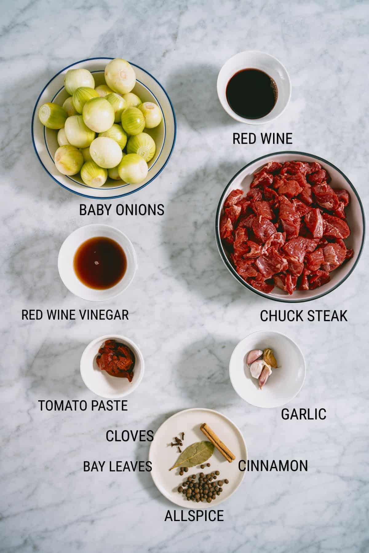Bowls on a marble surface containing labelled ingredients: baby onions, chuck steak, garlic, cinnamon, allspice, bay leaves, cloves, tomato purée, red wine vinegar, and red wine.