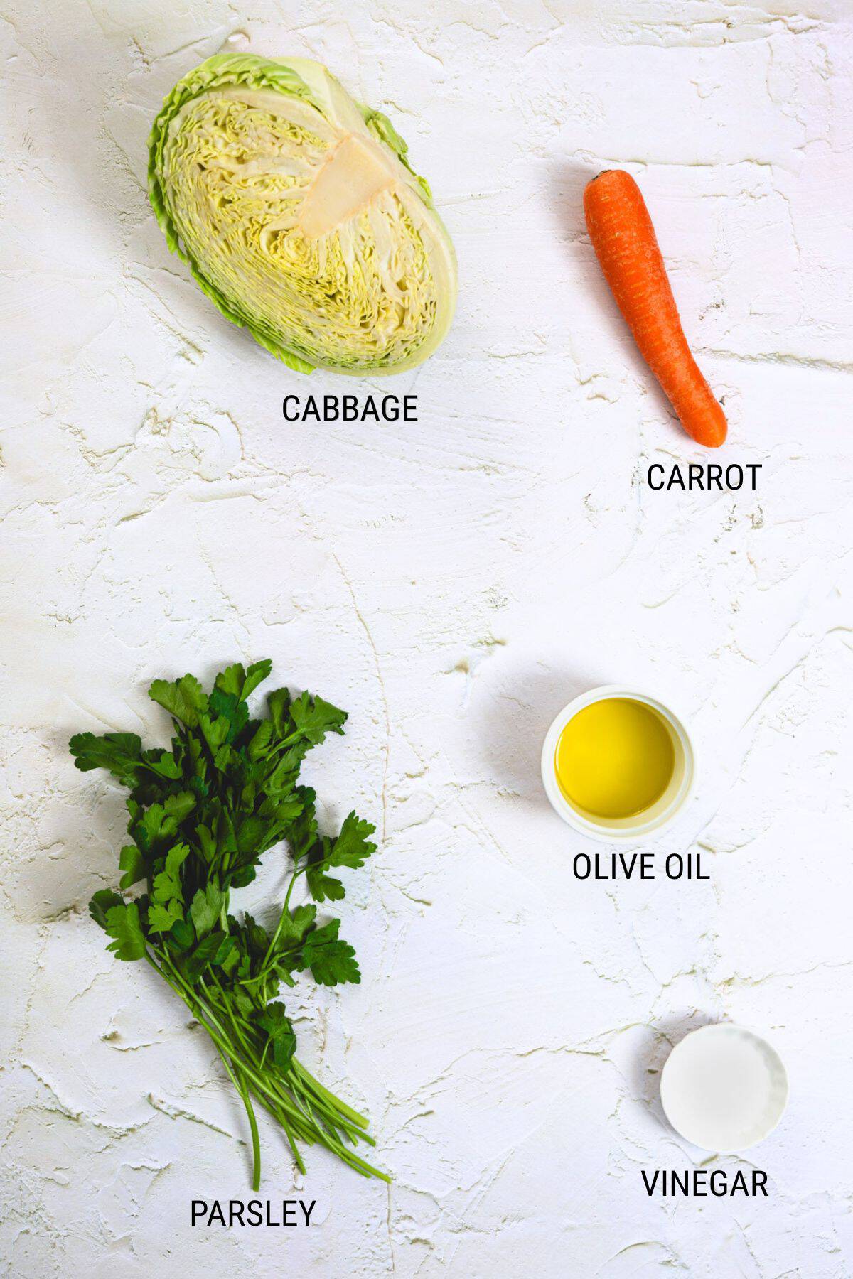 Half a cabbage, a whole carrot, parsley, olive oil, and vinegar are arranged on a white textured surface and labelled-perfect fresh ingredients for making a vibrant Greek cabbage salad.