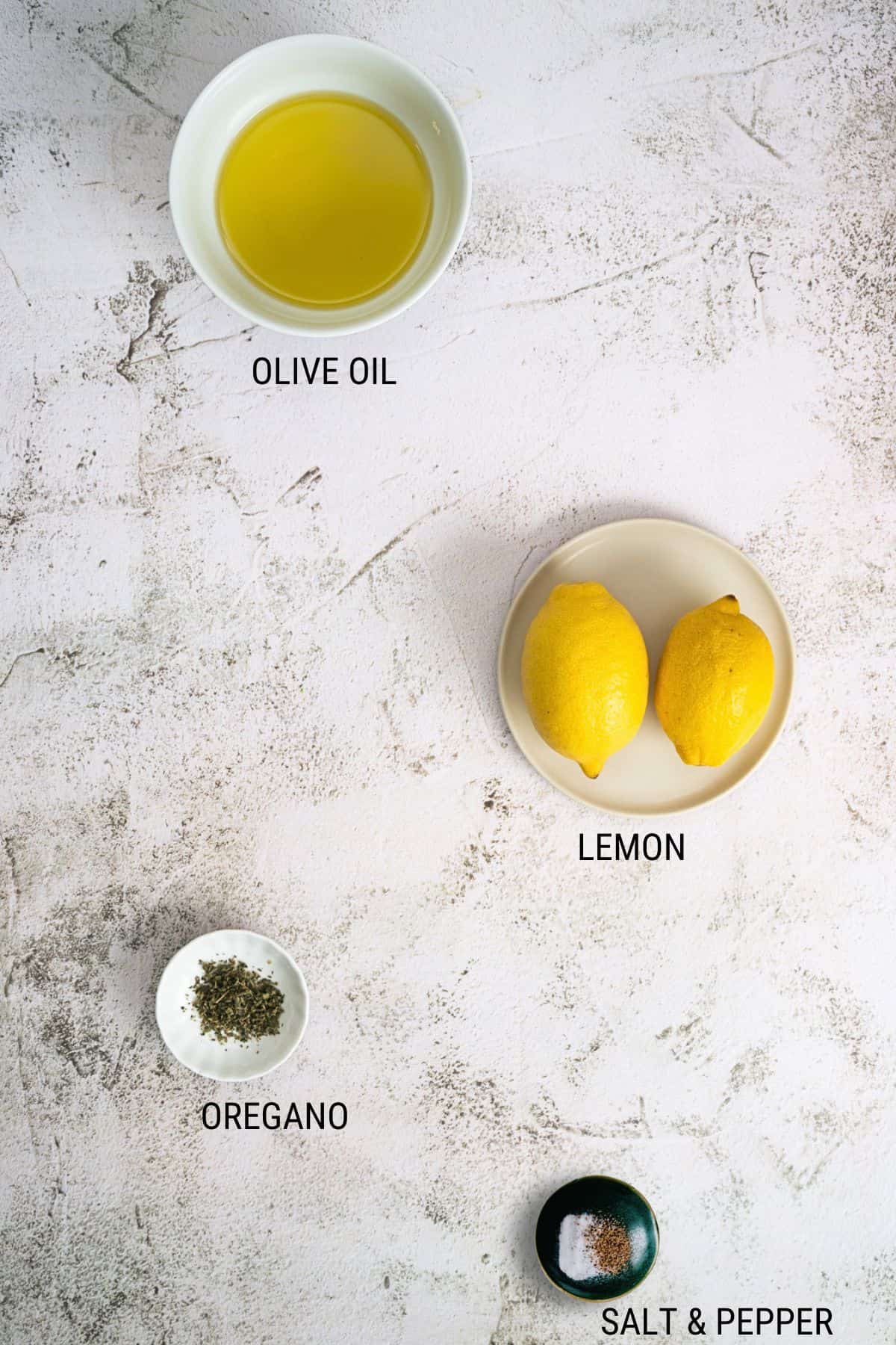 Flat lay of ingredients on a white surface: a bowl of olive oil, two whole lemons on a small plate, a small dish of dried oregano, and a small bowl of salt and pepper. Each item is labelled.