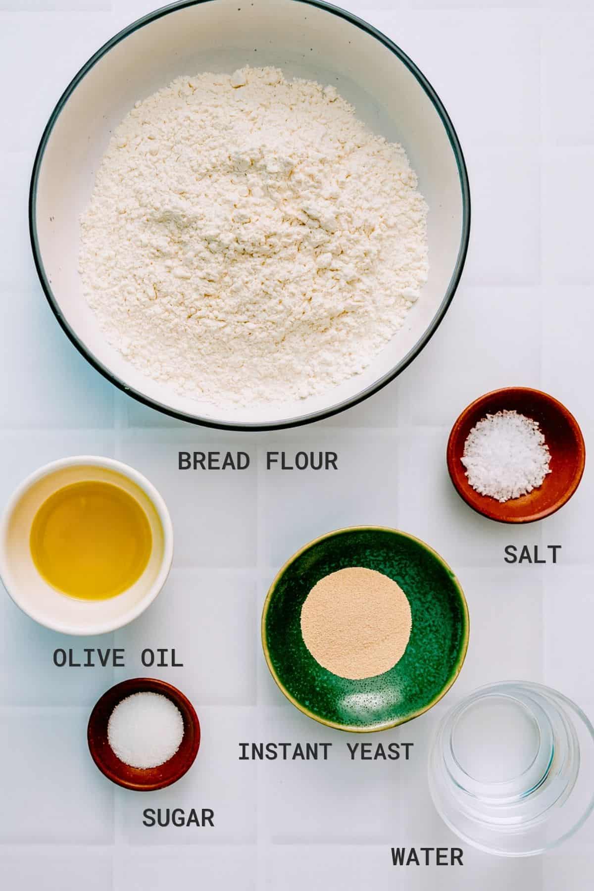 A bowl of bread flour surrounded by small bowls of salt, olive oil, instant yeast, sugar, and a glass of water, all labelled with their names, arranged on a white tiled surface.