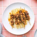 Five skewers of grilled marinated pork souvlaki are arranged on a white plate with some sauce, placed on a pink tiled surface. A striped cloth is partially visible in the bottom left corner.