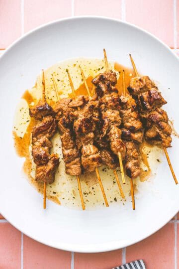 Five skewers of grilled marinated pork souvlaki are arranged on a white plate with some sauce, placed on a pink tiled surface. A striped cloth is partially visible in the bottom left corner.