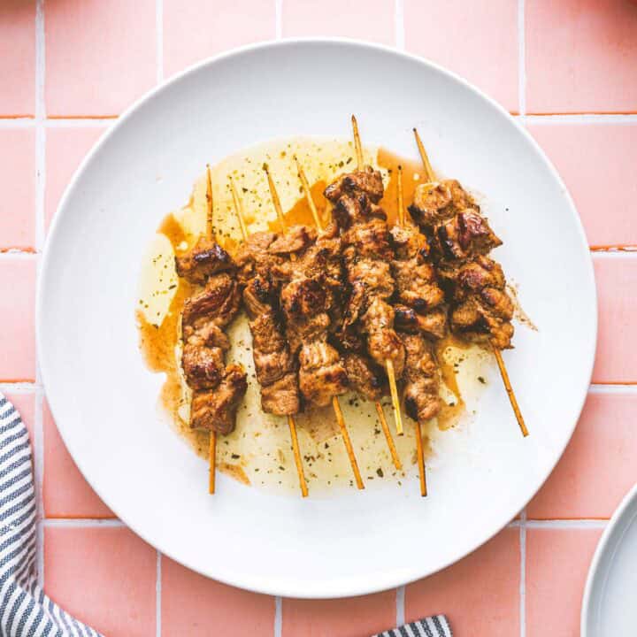 Five skewers of grilled marinated pork souvlaki are arranged on a white plate with some sauce, placed on a pink tiled surface. A striped cloth is partially visible in the bottom left corner.
