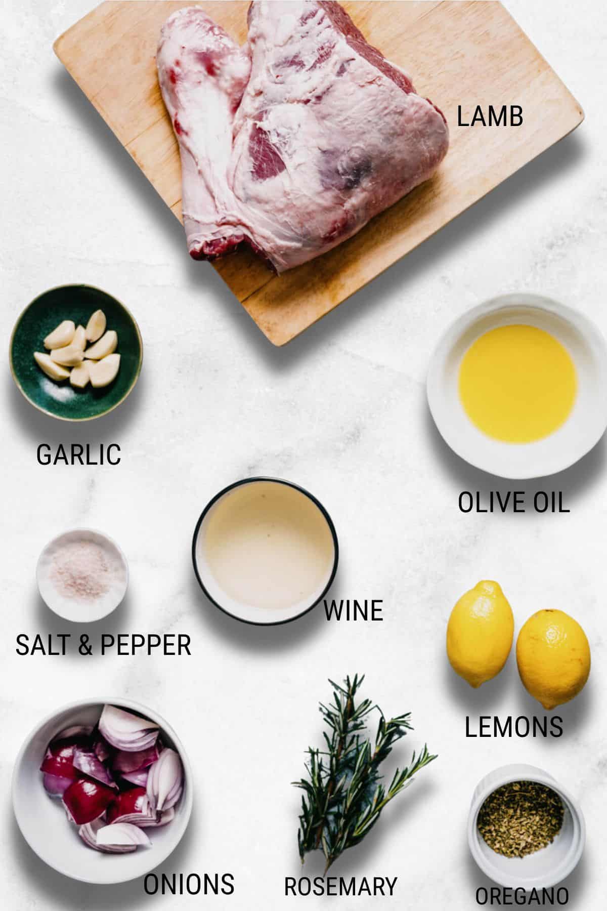 Raw lamb on a wooden board with labelled ingredients nearby: garlic cloves, olive oil, salt and pepper, wine, lemons, red onion pieces, fresh rosemary, and dried oregano on a light background.