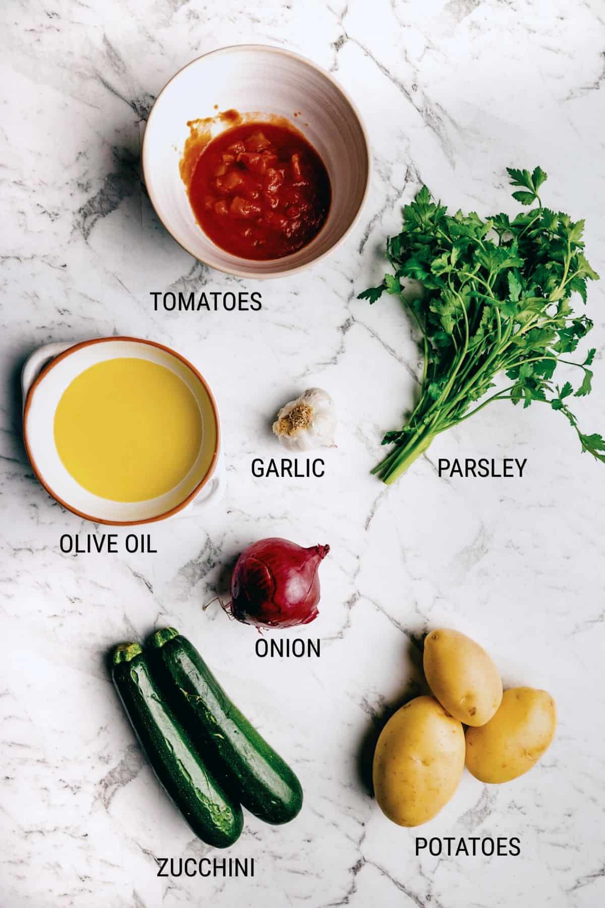 A marble surface displays bowls of chopped tomatoes and olive oil, along with garlic, parsley, a red onion, two courgettes, and three yellow potatoes. Each ingredient is labelled with black text.