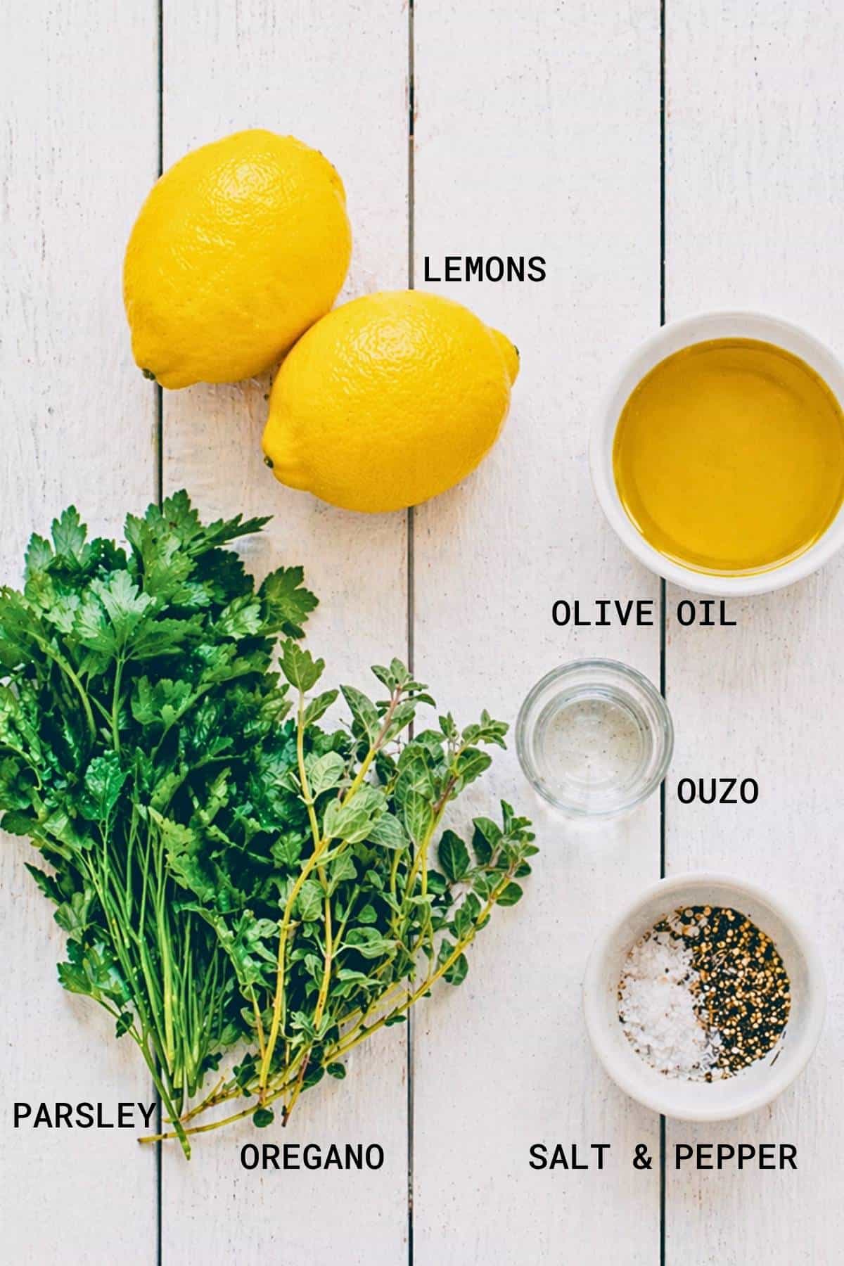 Overhead view of fresh parsley, oregano, two lemons, a bowl of olive oil, a small glass of ouzo, and a bowl containing salt and pepper, all labelled on a white wooden surface.