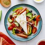 A blue plate with Greek salad featuring sliced tomatoes, cucumbers, red onions, peppers, black olives, and a large wedge of feta cheese sprinkled with herbs. A wooden spoon with dried oregano is nearby.