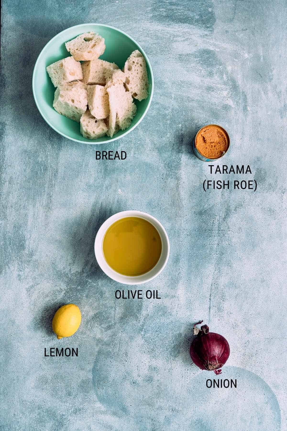 A flat lay of ingredients labelled: bread in a bowl, tarama (fish roe), a bowl of olive oil, a lemon, and a red onion on a textured blue surface.