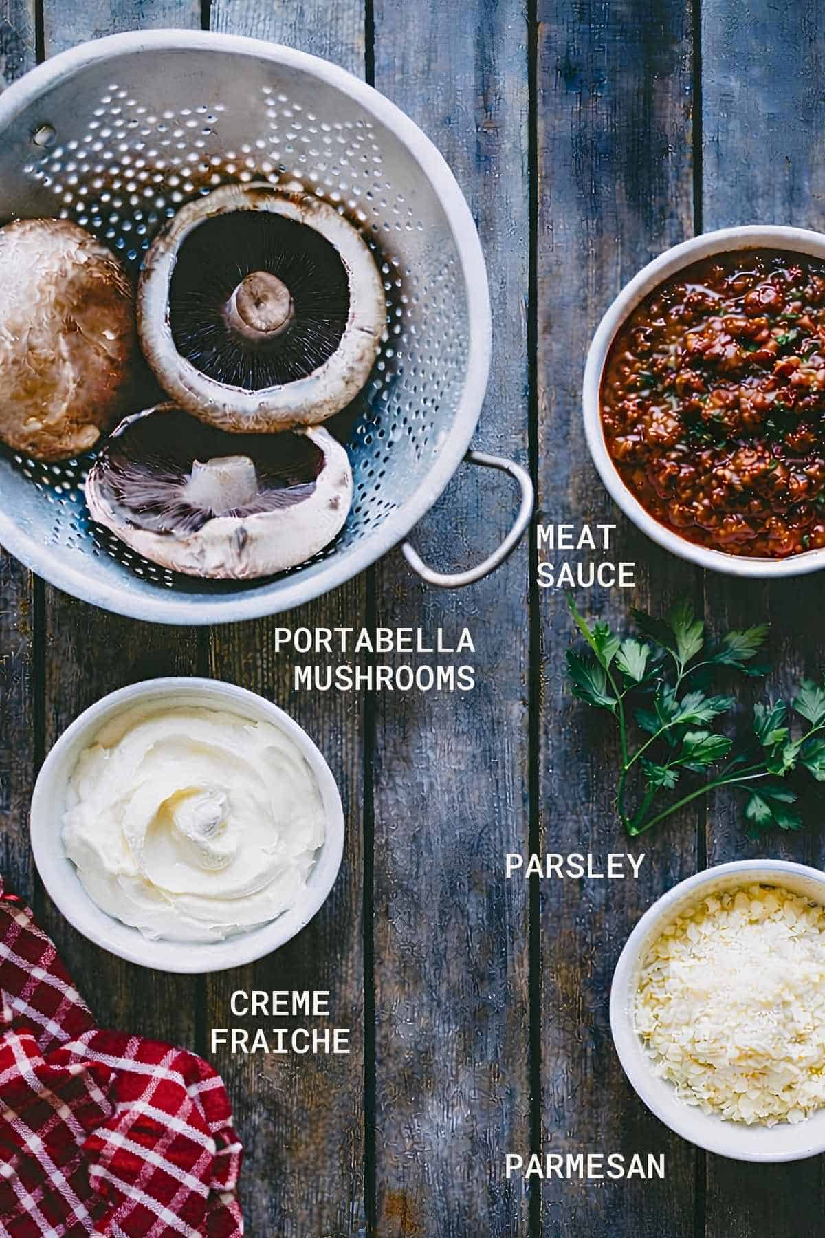 Overhead view of ingredients on a wooden table: portobello mushrooms in a colander, a bowl of meat sauce, parsley, a bowl of crème fraîche, and a bowl of grated Parmesan cheese, with labels for each.