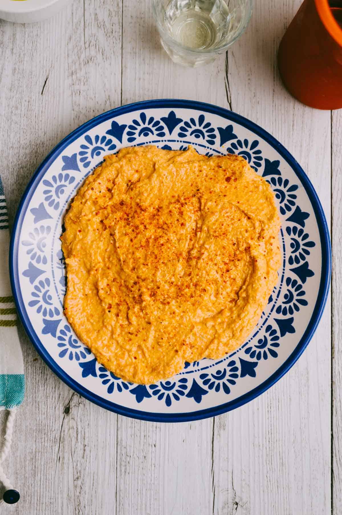 A blue and white patterned plate with a spread of spicy orange htipiti dip, sprinkled with spices, sits on a white wooden table next to a glass of water and a striped cloth.