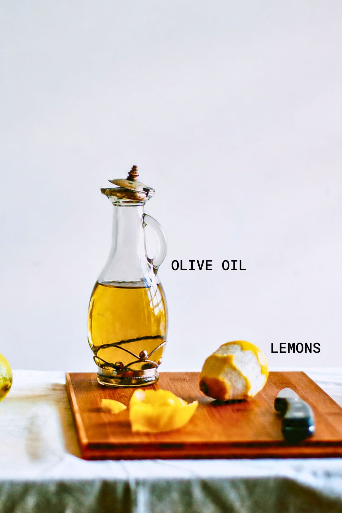 A glass bottle of olive oil stands on a wooden board next to a partially peeled lemon, lemon peel, and a small knife. Text labels identify the olive oil and lemons. The scene is set on a light background.