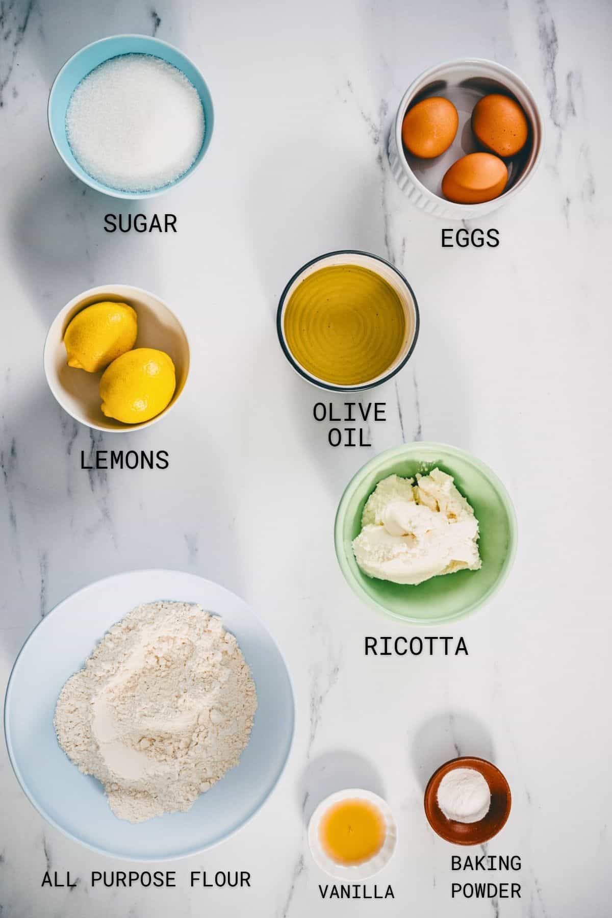 Flat lay of baking ingredients on a white surface, each labelled: sugar, eggs, lemons, olive oil, ricotta, plain flour, vanilla, and baking powder, each ingredient in a separate bowl or container.