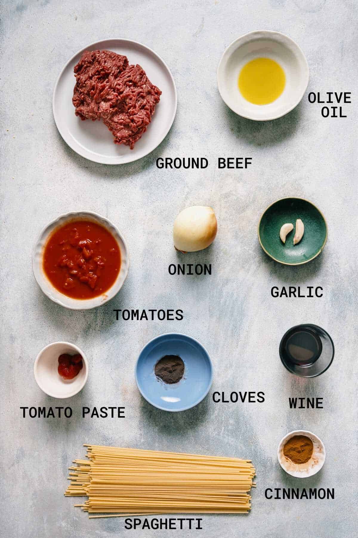 A flat lay of labelled ingredients for spaghetti: minced beef, olive oil, onion, garlic, tomatoes, tomato purée, cloves, wine, cinnamon, and uncooked spaghetti, all arranged on a light surface.