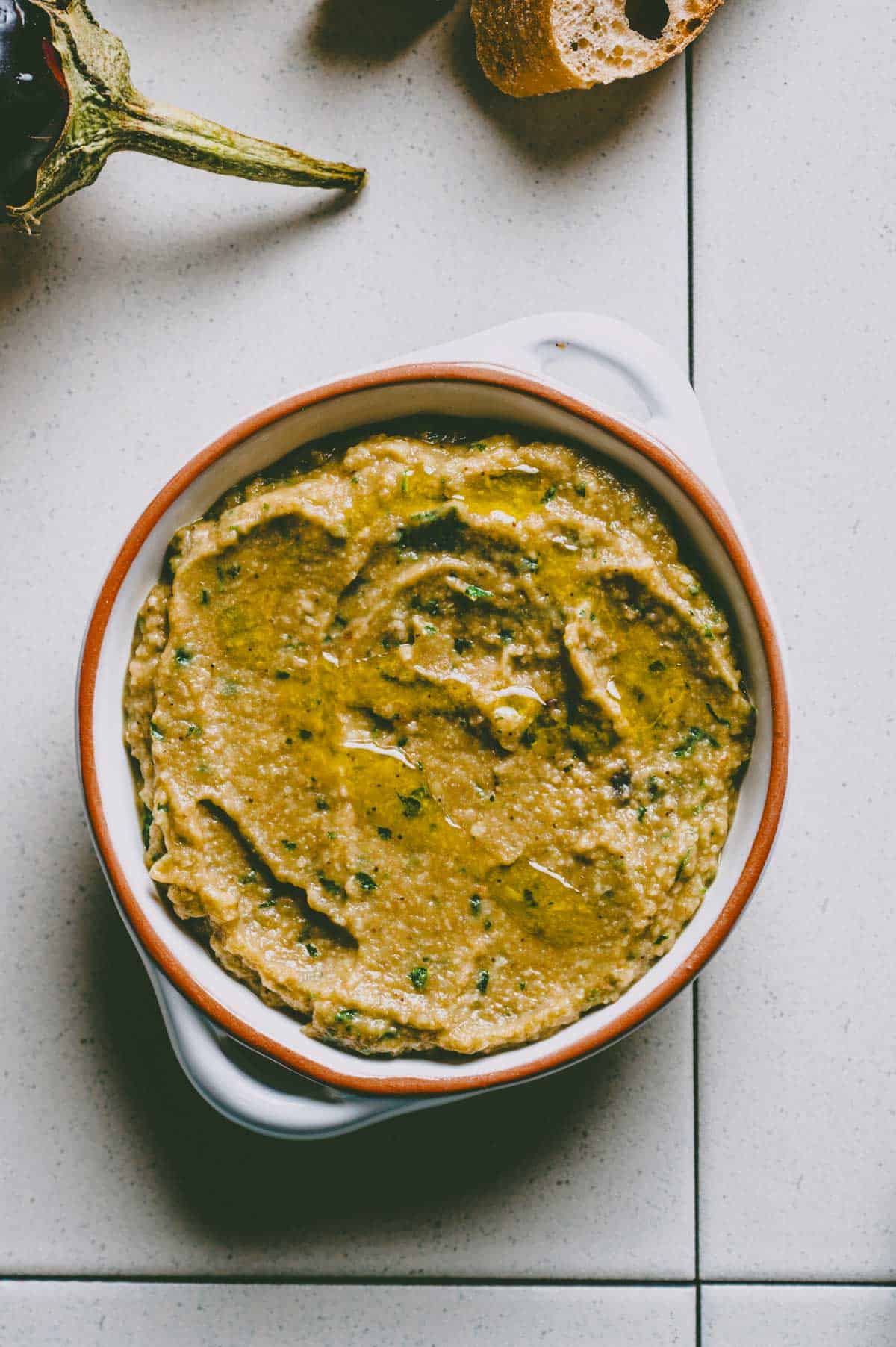 A bowl of creamy greek eggplant dip garnished with herbs and a drizzle of olive oil, placed on a tiled surface with a piece of bread and part of an aubergine nearby.