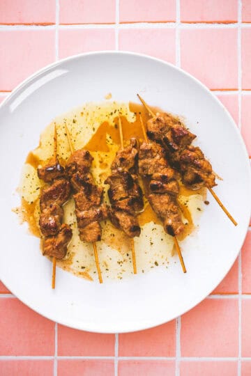 A white plate with five grilled pork souvlaki drizzled with sauce, placed on a pink tiled surface.
