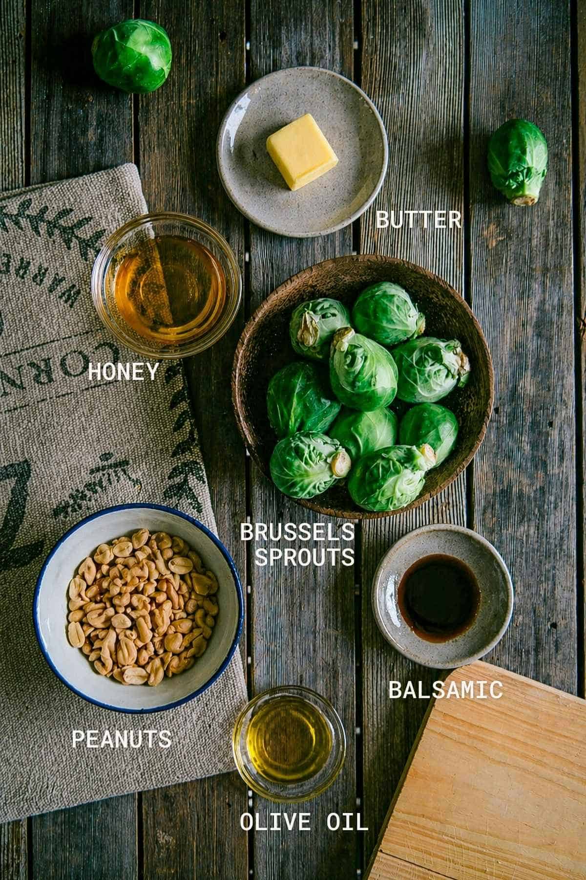 Overhead view of ingredients on a wooden table: a bowl of Brussels sprouts, butter on a plate, small bowls of honey, balsamic vinegar, peanuts, and olive oil, each labelled with white text.