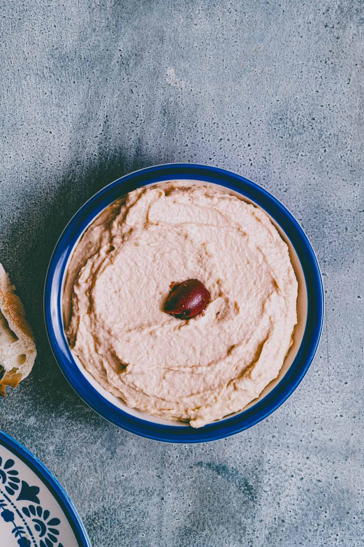 A bowl of creamy taramasalata topped with a single black olive, placed on a grey textured surface next to a piece of bread and a partial blue-and-white patterned plate.