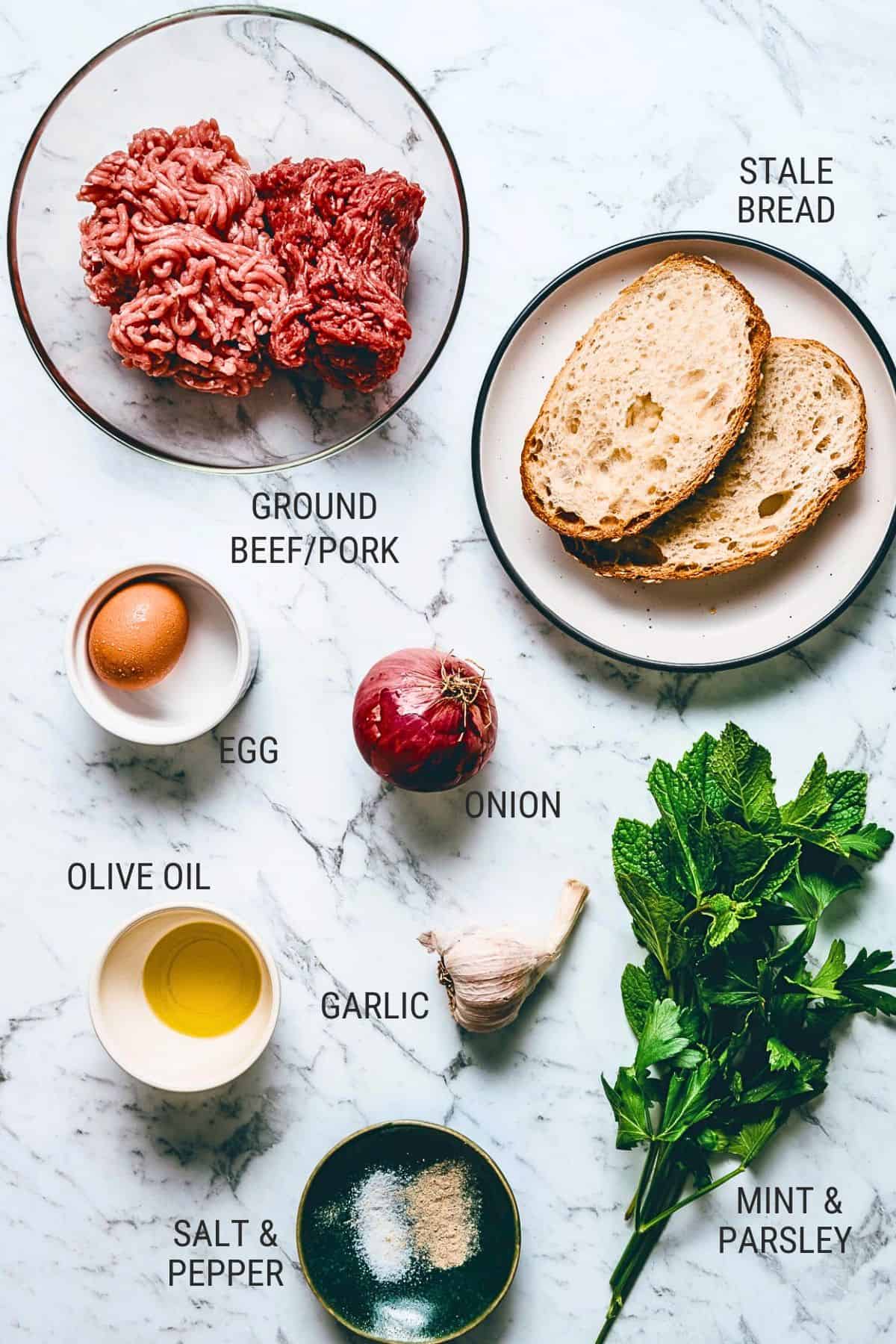 A top view of ingredients on a marble surface: a bowl of minced beef and pork, slices of stale bread, an egg, a red onion, garlic, olive oil, salt and pepper, and fresh mint and parsley. Each ingredient is labelled.