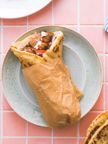 A wrapped gyros with grilled meat, tomatoes, and sauce served on a speckled plate over a pink tiled surface, with a striped serviette and pieces of pitta bread nearby.
