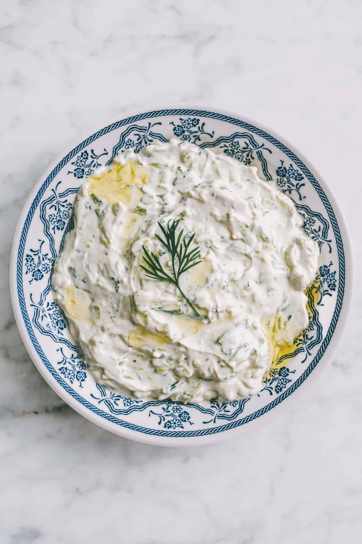 A blue and white patterned bowl filled with creamy tzatziki sauce, garnished with a sprig of dill and a drizzle of olive oil, on a white marble surface.