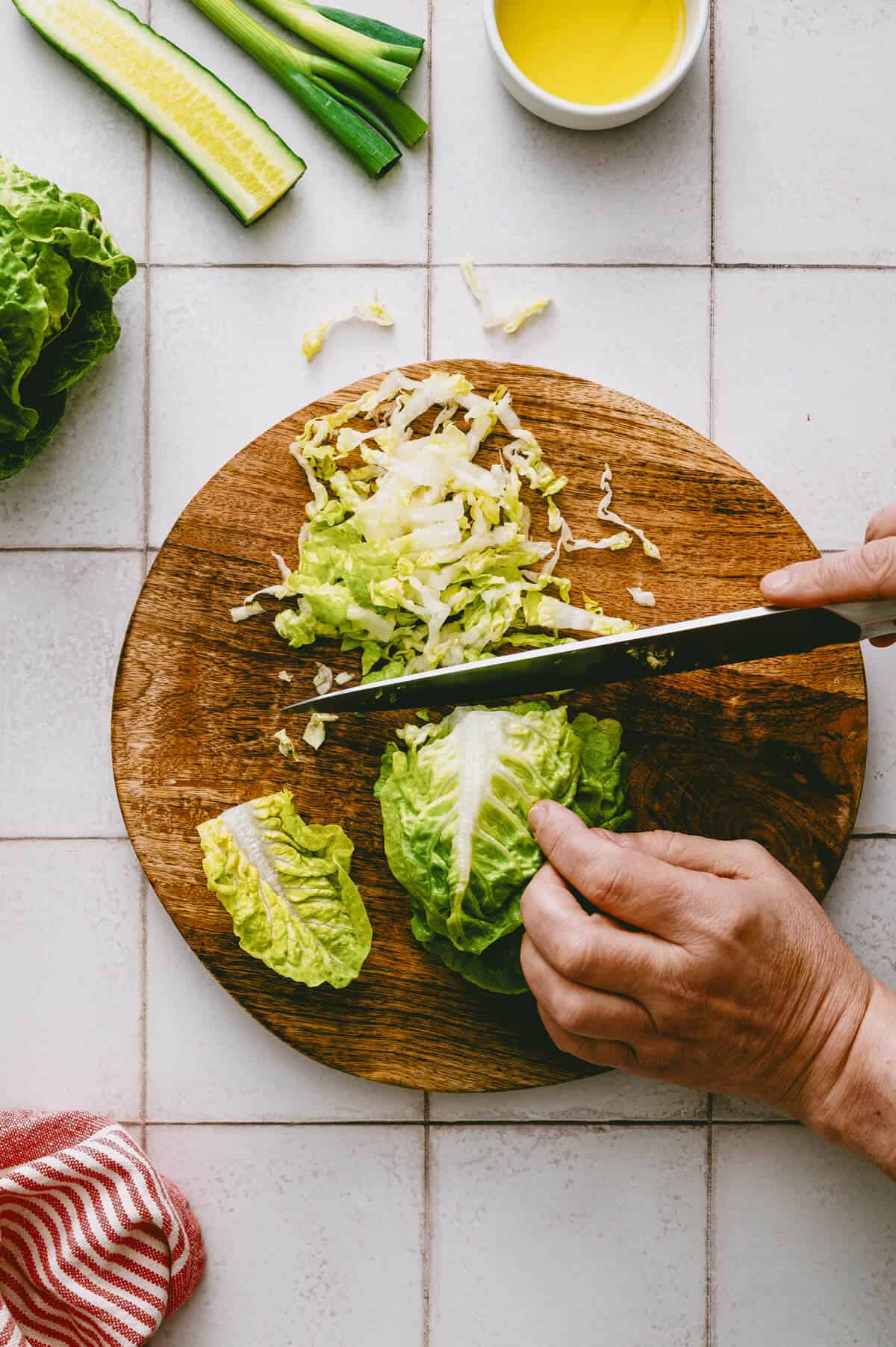 Hands slicing cos lettuce on a round wooden chopping board, surrounded by cucumber, spring onions, a cup of olive oil, and a red-striped tea towel on a tiled kitchen worktop.