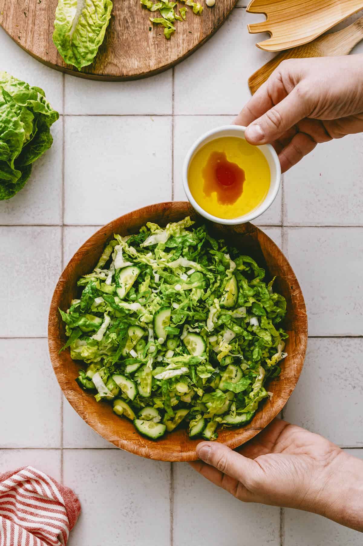 A person holds a small bowl of oil and vinegar above a wooden bowl filled with chopped lettuce and cucumber, preparing to dress the salad. Fresh lettuce and cutlery are nearby on a tiled surface.