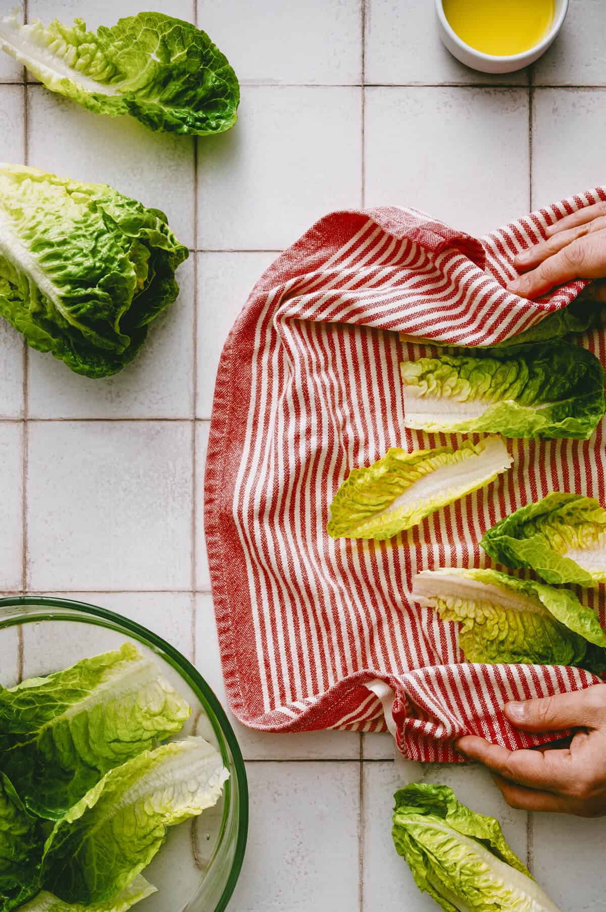 Romaine lettuce leaves being dried on a red and white striped tea towel, with a bowl of lettuce, a cup of yellow liquid, and lettuce leaves on a white tiled surface.