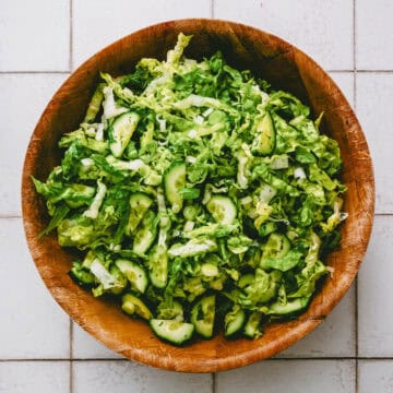 A wooden bowl filled with a fresh green salad made of chopped lettuce, cucumber slices, and spring onions, placed on a white tiled surface.