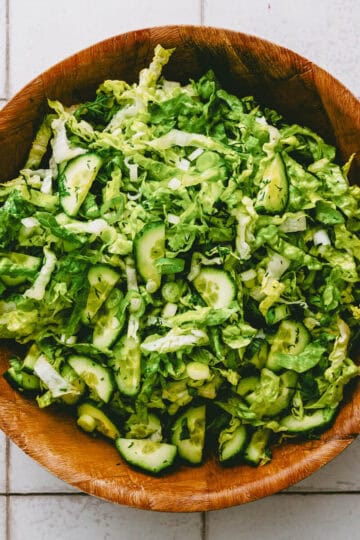 A wooden bowl filled with a fresh green salad made of chopped lettuce, cucumber slices, and spring onions, placed on a white tiled surface.