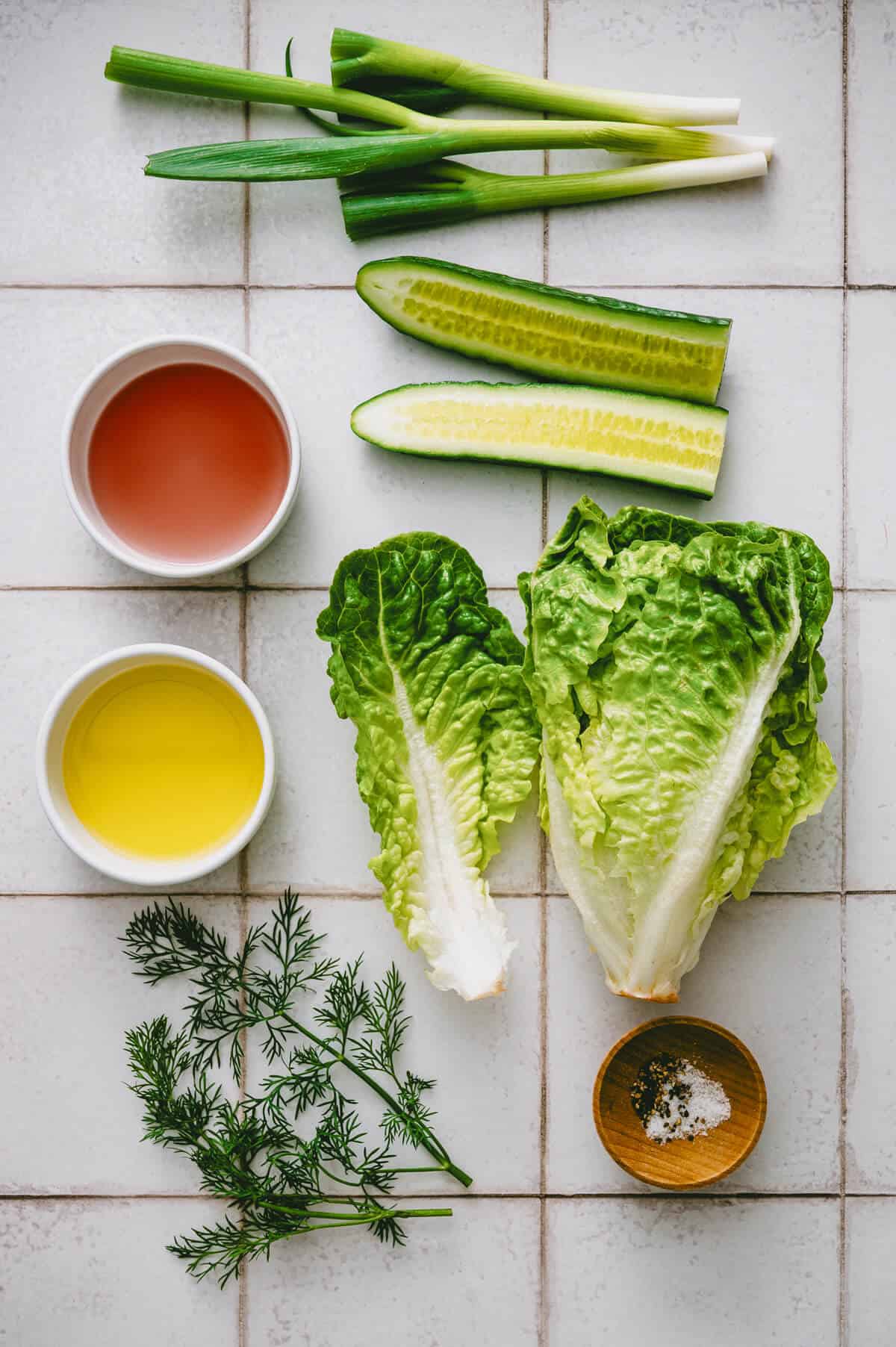 Fresh salad ingredients on a tiled surface: spring onions, cucumber, two romaine lettuce leaves, dill, a small dish of salt and pepper, and two bowls containing olive oil and red wine vinegar.