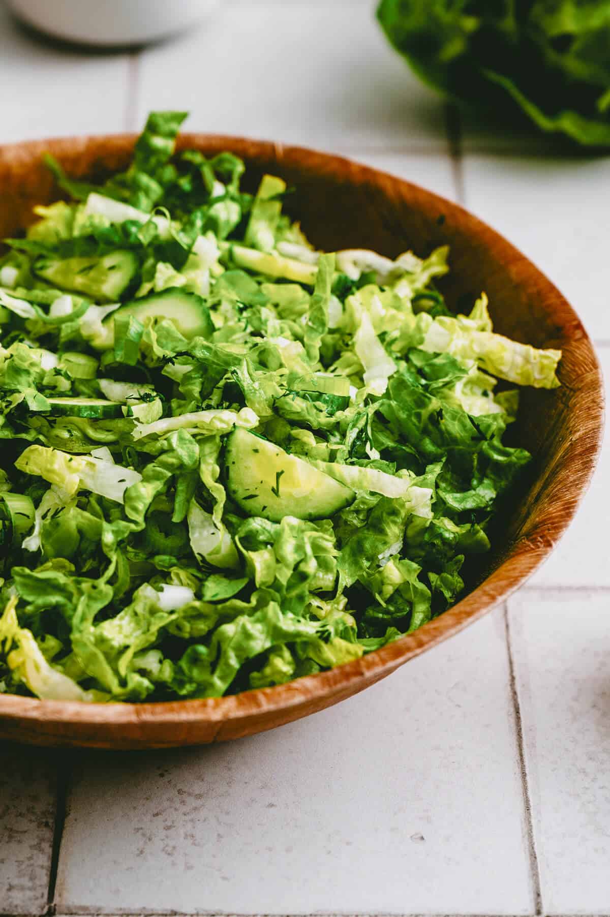 A wooden bowl filled with a fresh green salad, featuring chopped lettuce and cucumber slices, sits on a white tiled surface.