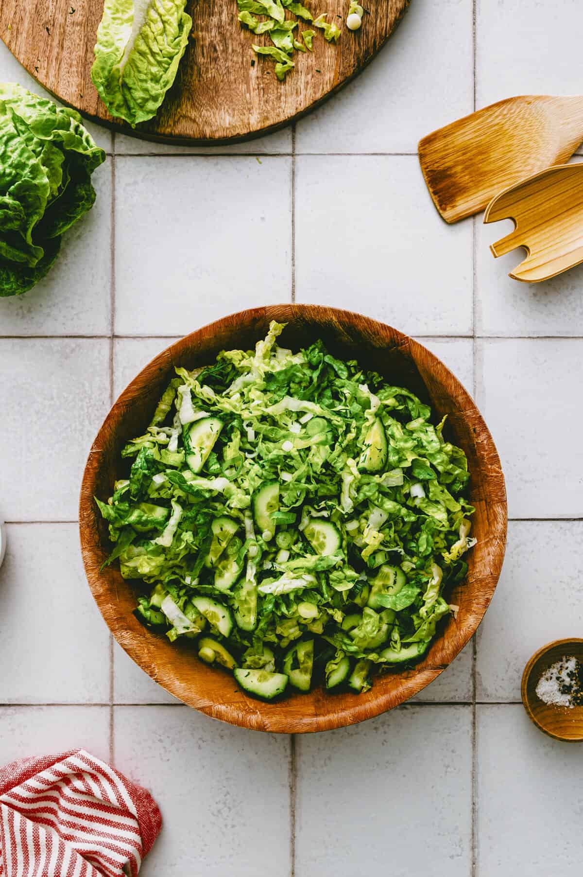 A wooden bowl filled with chopped green lettuce and cucumber slices sits on a tiled worktop, surrounded by a chopping board with lettuce, wooden salad servers, a small bowl of seasoning, and a red-striped tea towel.