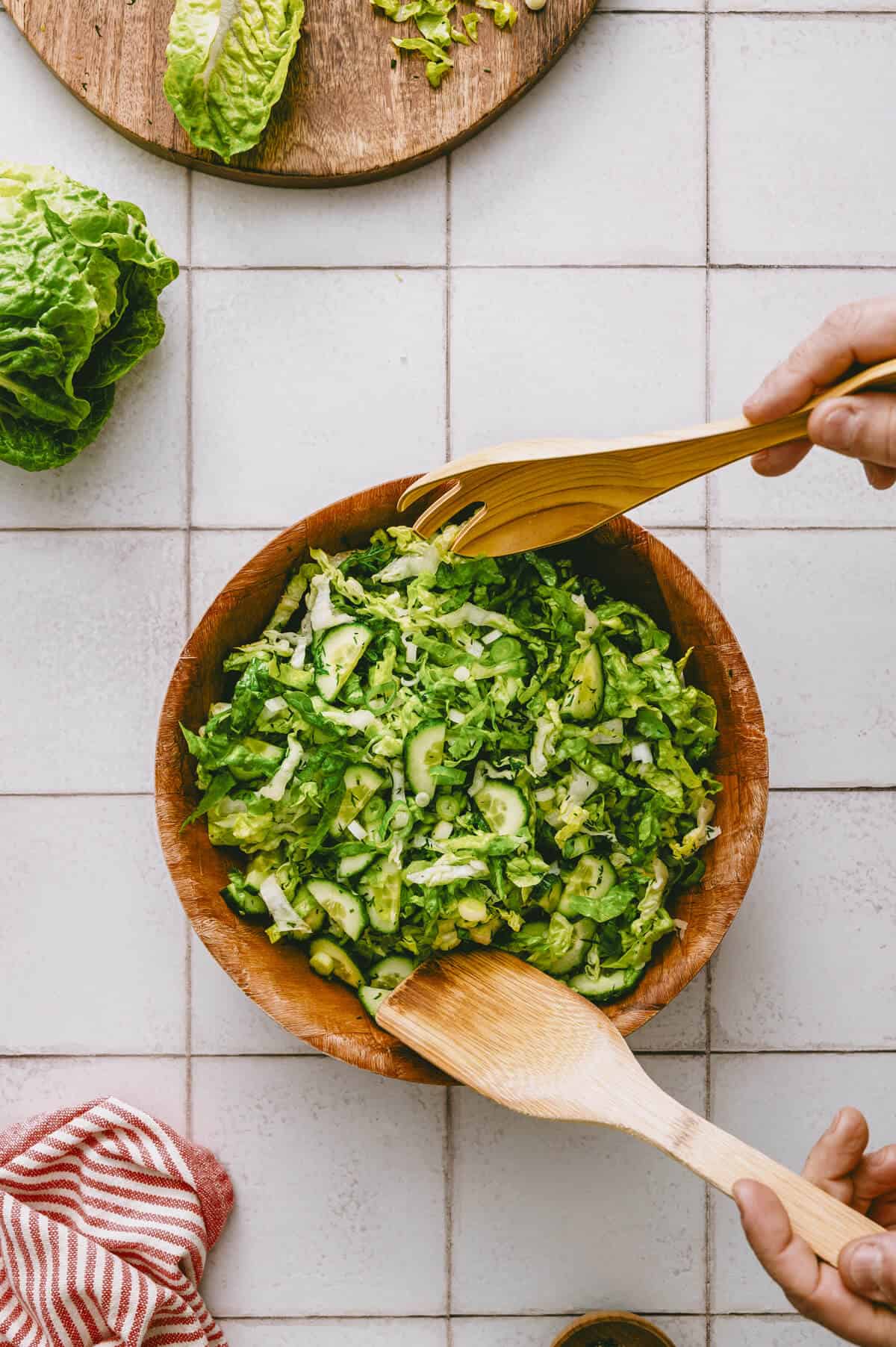 A hand tosses a fresh green salad with cucumber and lettuce in a wooden bowl using wooden salad servers. Lettuce and a striped tea towel are nearby on a white tiled surface.