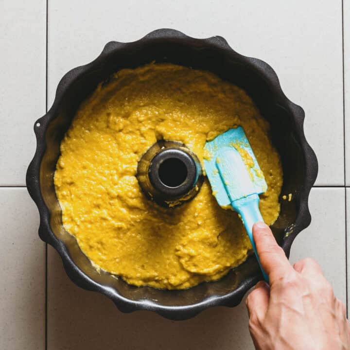 A hand uses a blue spatula to spread yellow cake mixture evenly in a black bundt tin, which sits on a white tiled surface.