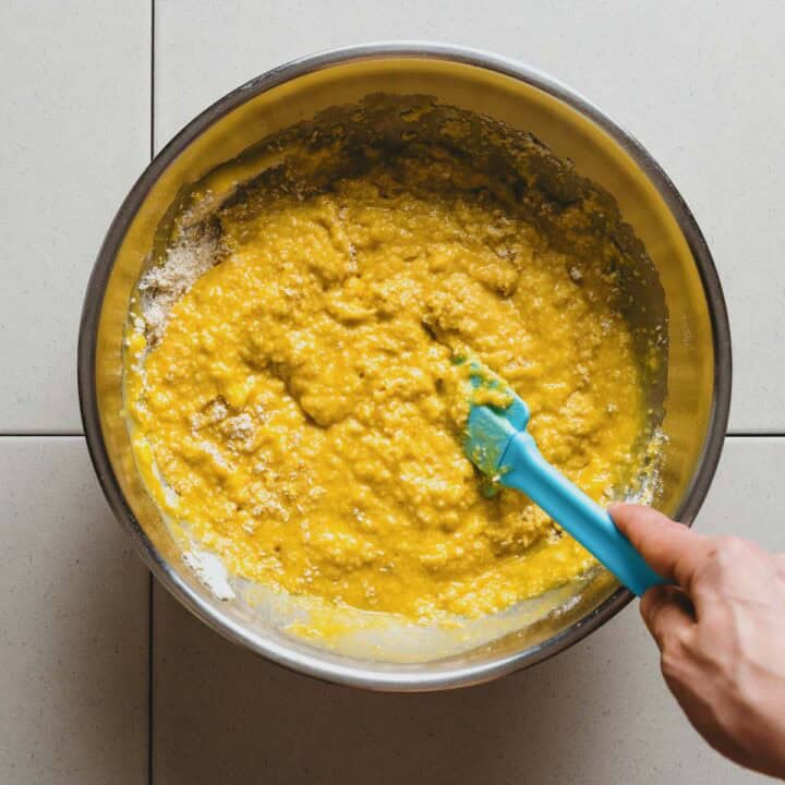 A hand is mixing yellow batter with a blue spatula in a large metal bowl placed on a light-coloured tiled surface.