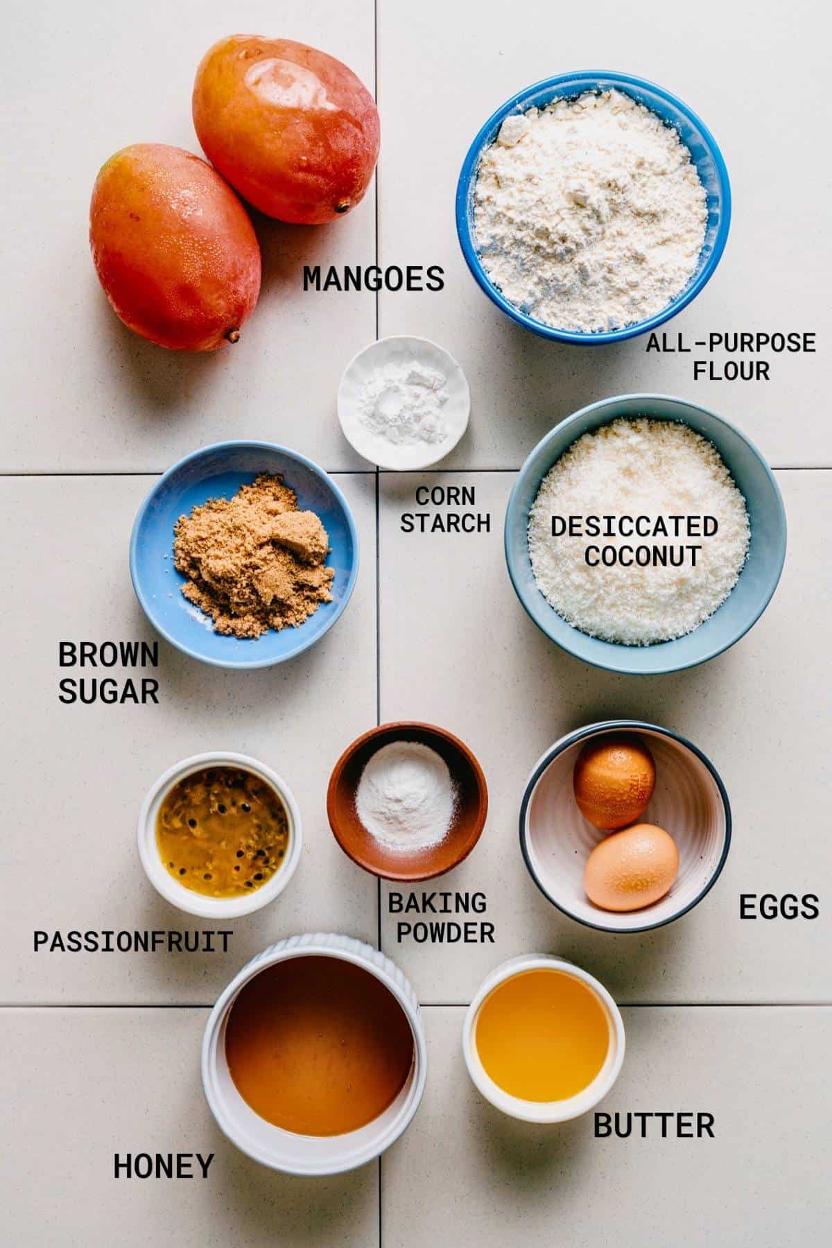Top-down view of labelled baking ingredients on a white surface: mangoes, plain flour, brown sugar, cornflour, desiccated coconut, passion fruit, baking powder, eggs, honey, and melted butter in bowls.