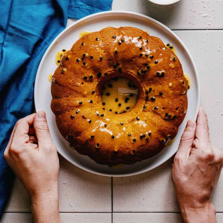 A person holds a white plate with a round bundt cake topped with passion fruit glaze and seeds, set on a tiled surface next to a blue serviette.