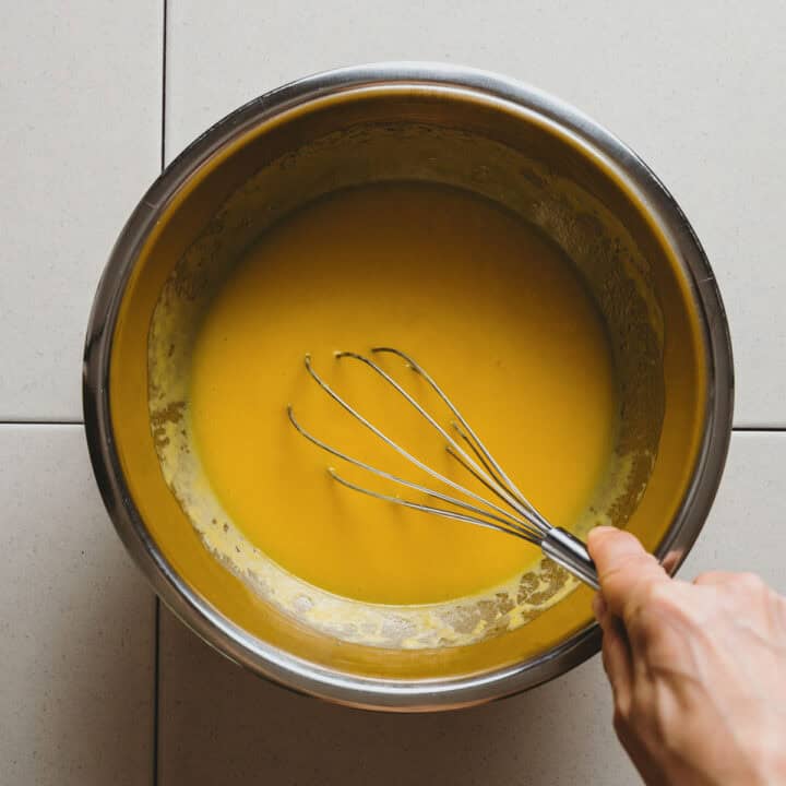 A hand holds a metal whisk, mixing a smooth yellow batter in a stainless steel bowl, set on a white tiled worktop.