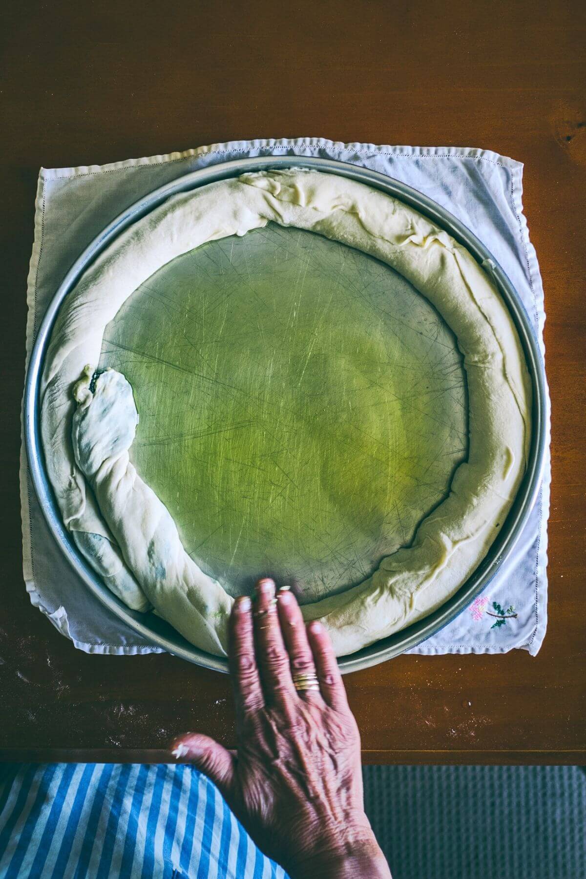 A person's hand pressing a coiled roll of dough into a round metal baking tin.