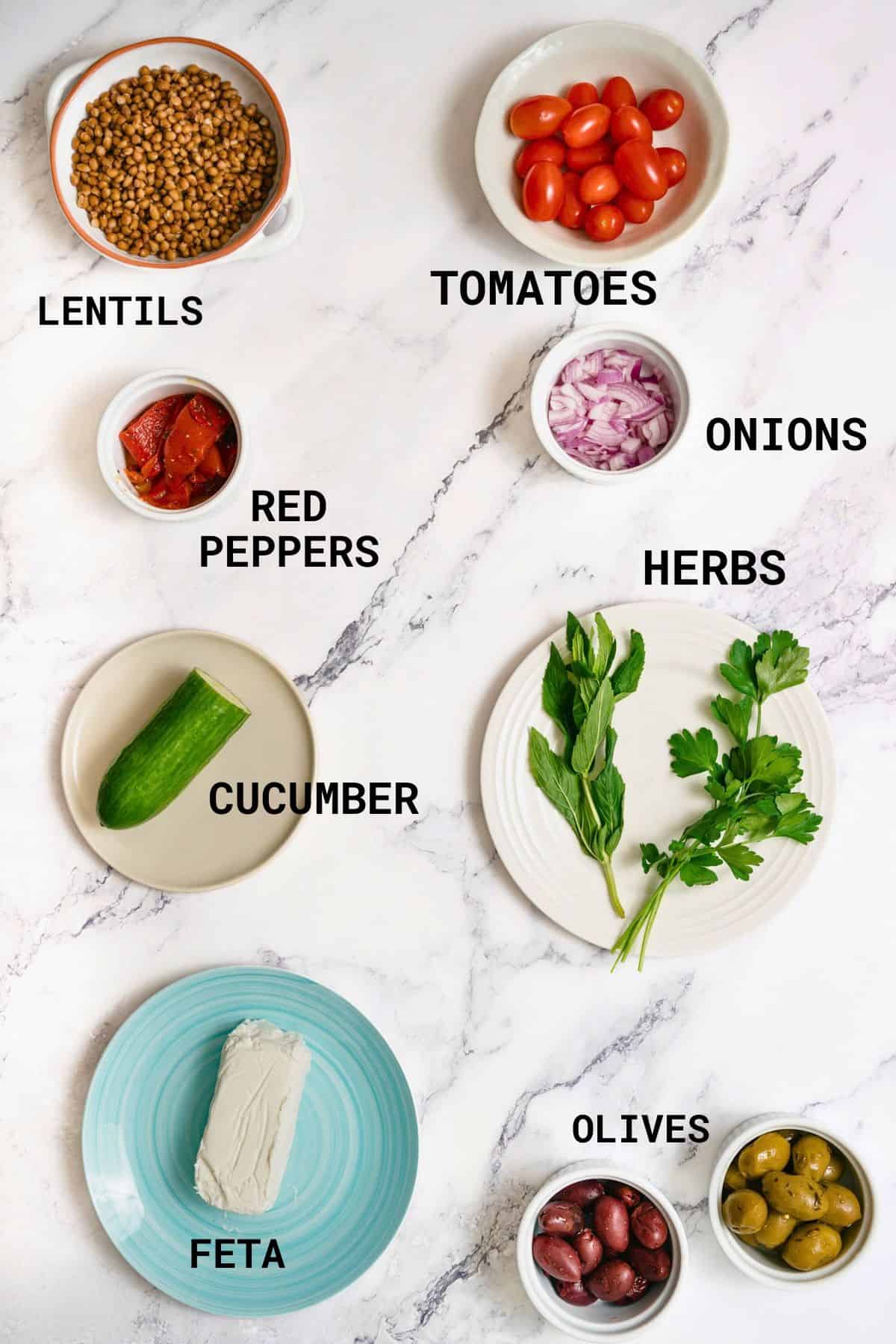 Ingredients for a Greek lentil salad laid out on a marble surface: lentils, cherry tomatoes, red peppers, onions, a cucumber, fresh herbs, a block of feta, and a mix of olives, with each item labelled.